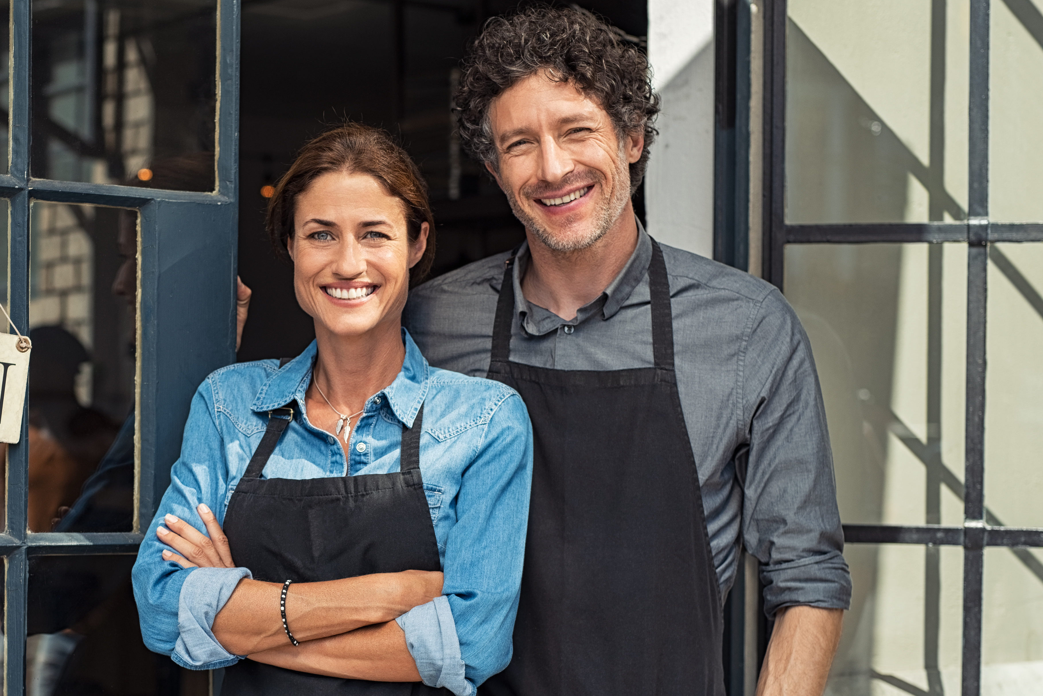 Two cheerful small business owners smiling and looking at camera while standing at entrance door. Happy mature man and mid woman at entrance of newly opened restaurant with open sign board. Smiling couple welcoming customers to small business shop.